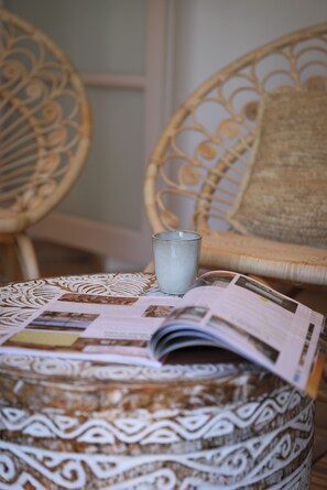 Una silla de mimbre con un cojín, un vaso de agua y una revista sobre un reposapiés estampado.