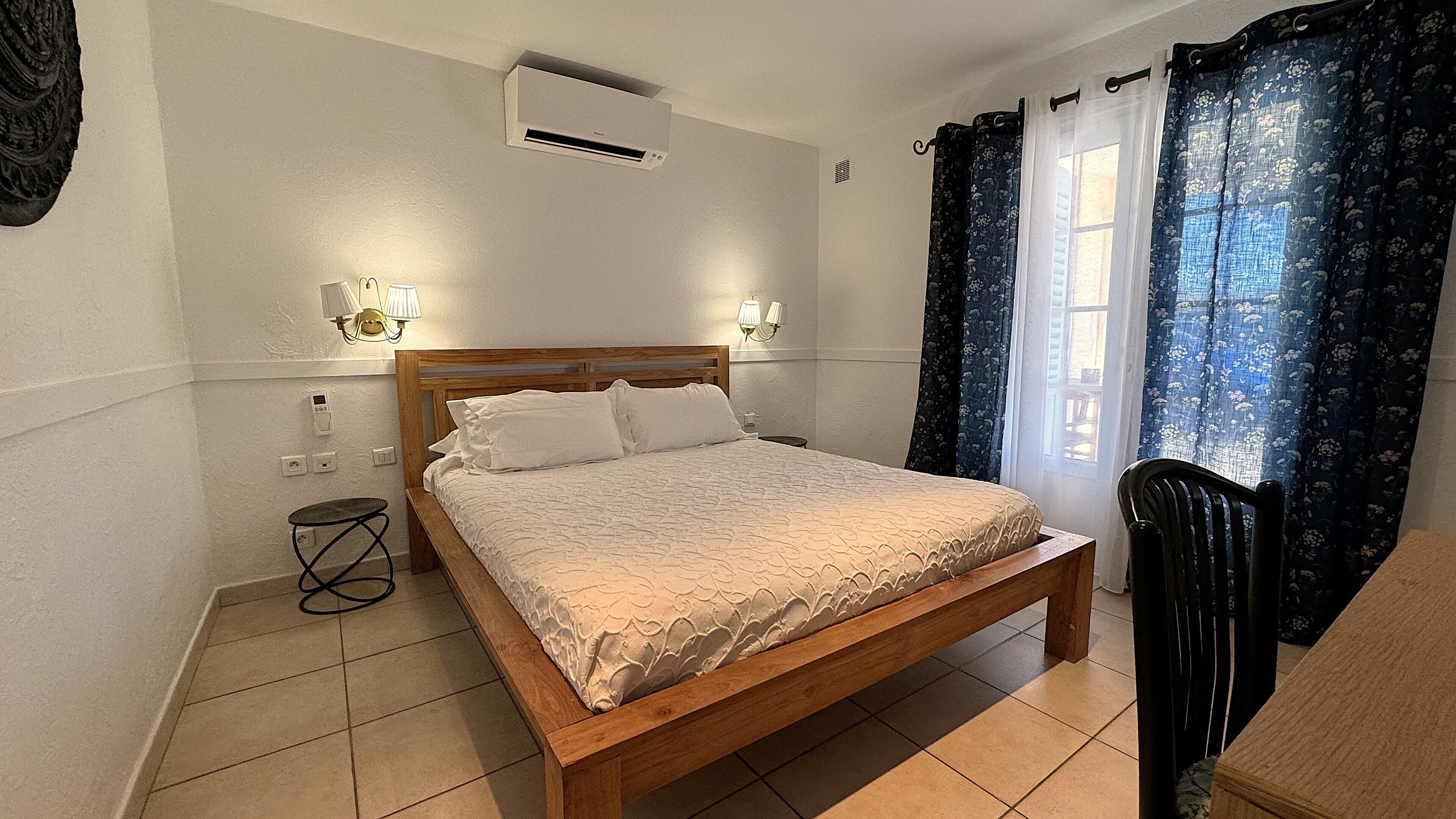 A bedroom with a wooden bed, two wall-mounted lamps, an air conditioning unit, and a window with floral curtains.