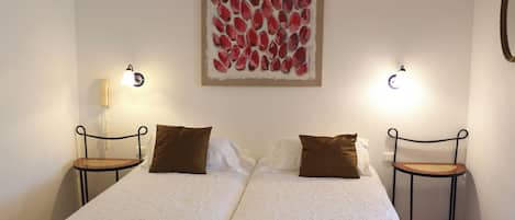 Een tweepersoonsbed met witte lakens, twee nachtkastjes met lampen, een kunstwerk aan de muur en een hanglamp.