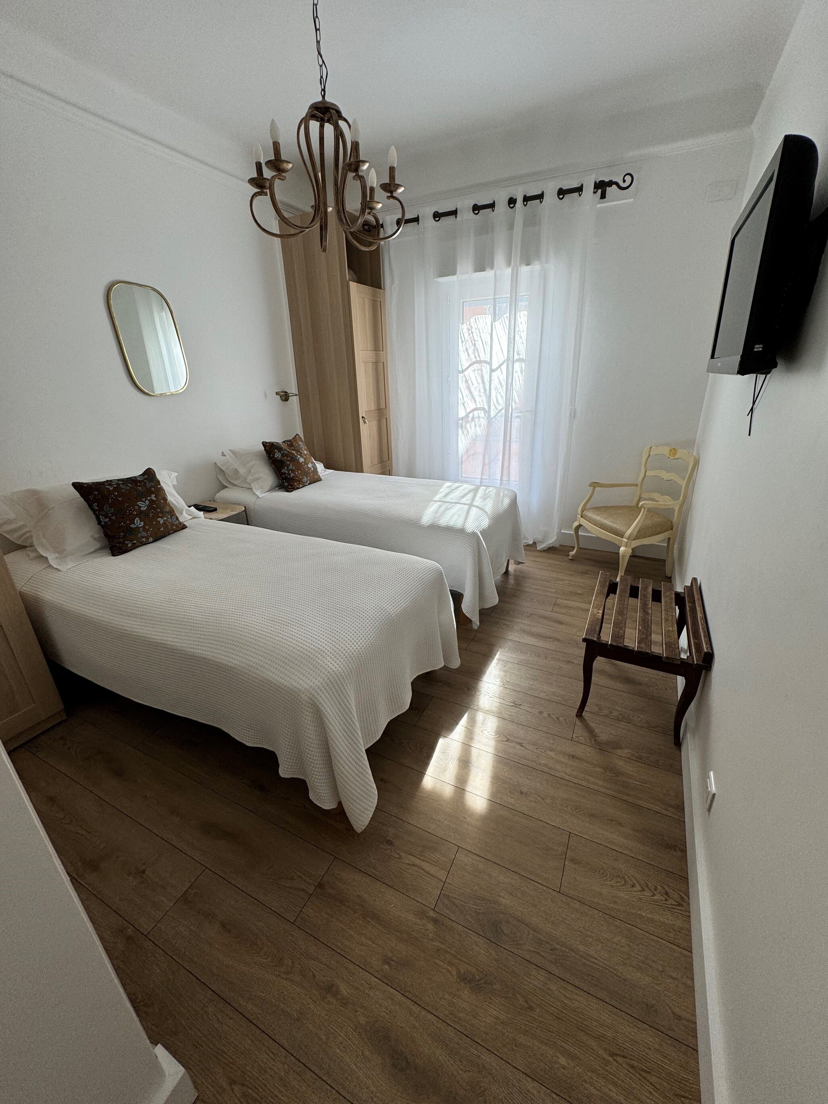 A room with two beds, a wooden floor, a chandelier, a chair, and a television on the wall.