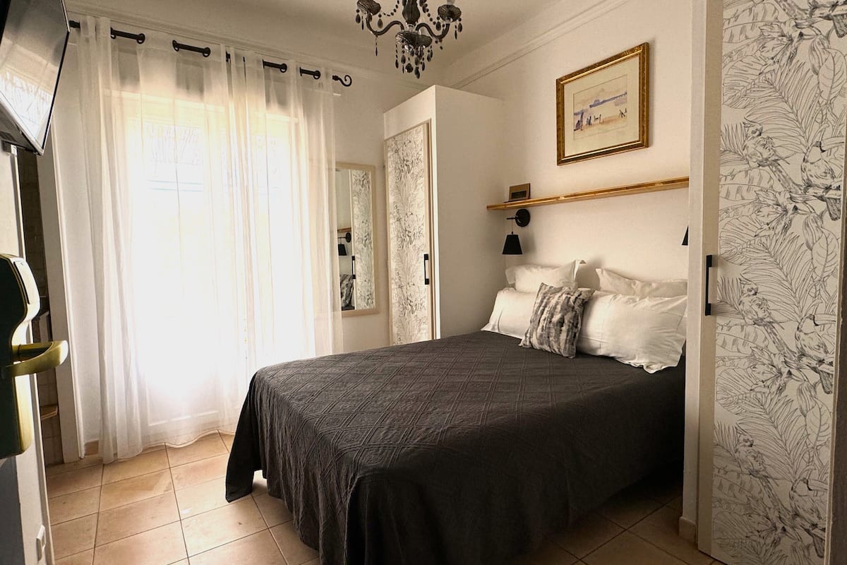 A bedroom with a bed, a bedside table, a mirror, and a window with curtains.