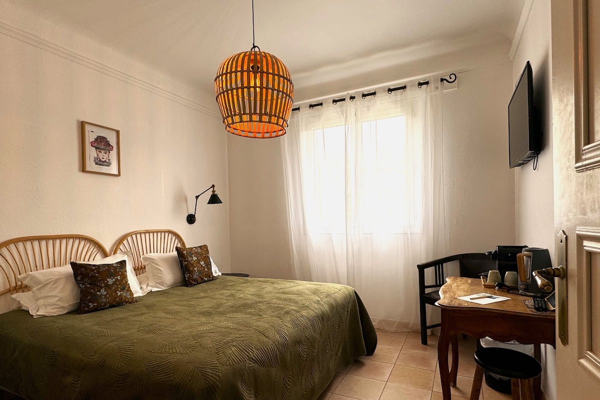 A bedroom with a bed, a desk, a chair, a lamp, a television, and a window with curtains.