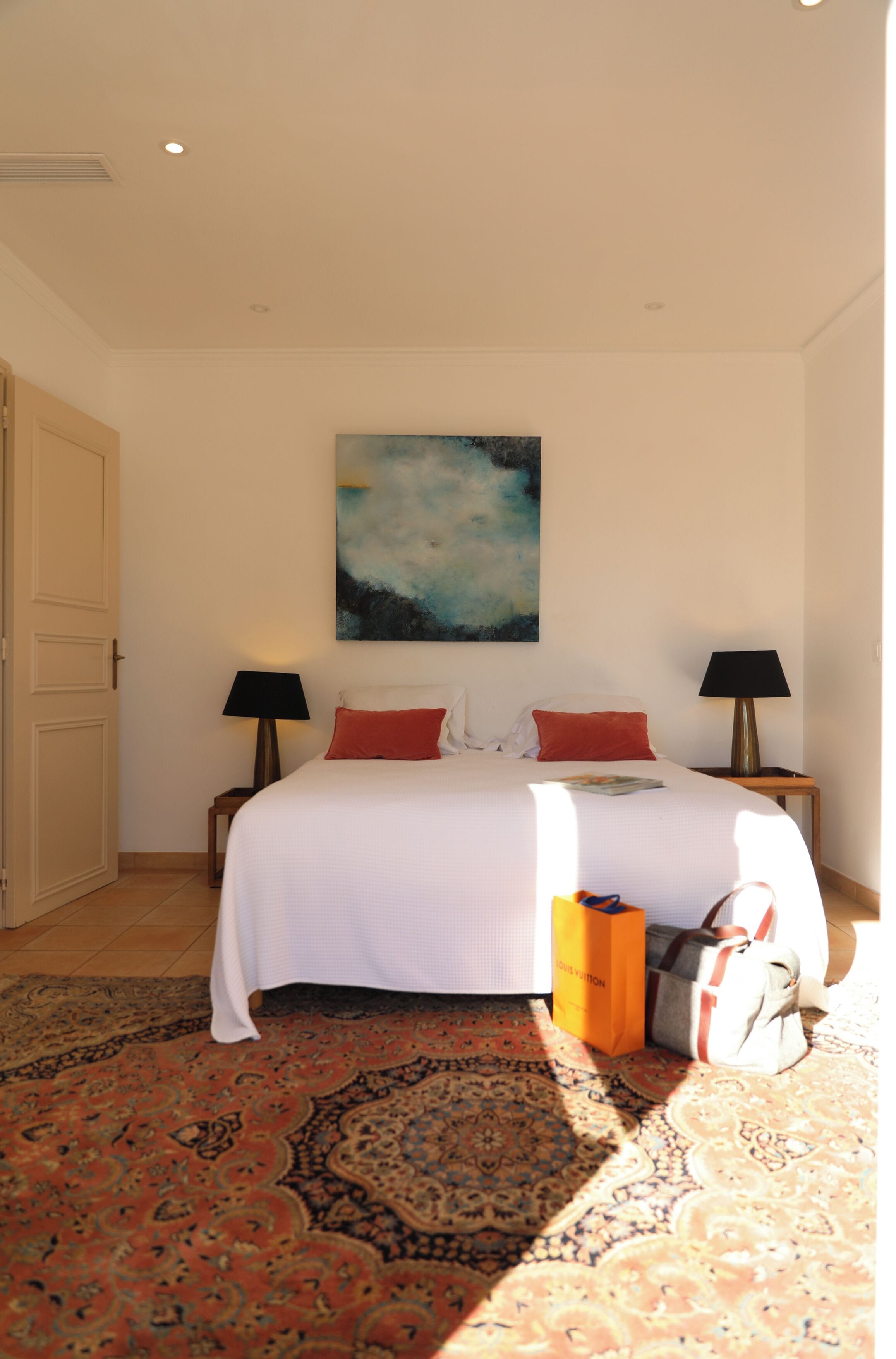 A hotel room with a large bed, two bedside tables, a painting on the wall, and a patterned rug.