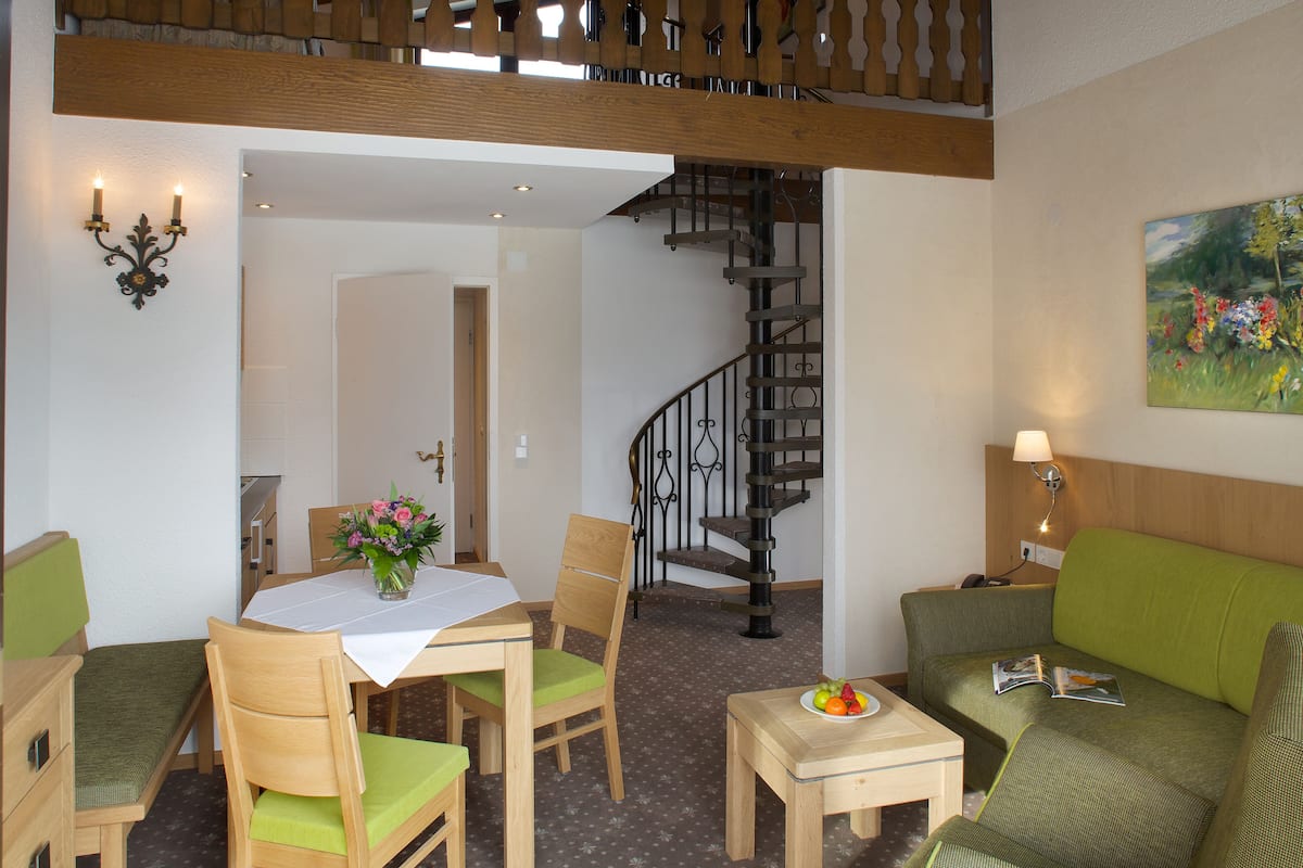 A dining area with a table, chairs, and a vase of flowers. There is a staircase with a railing leading to another room. A green sofa and a small table with a plate of fruit are also visible.