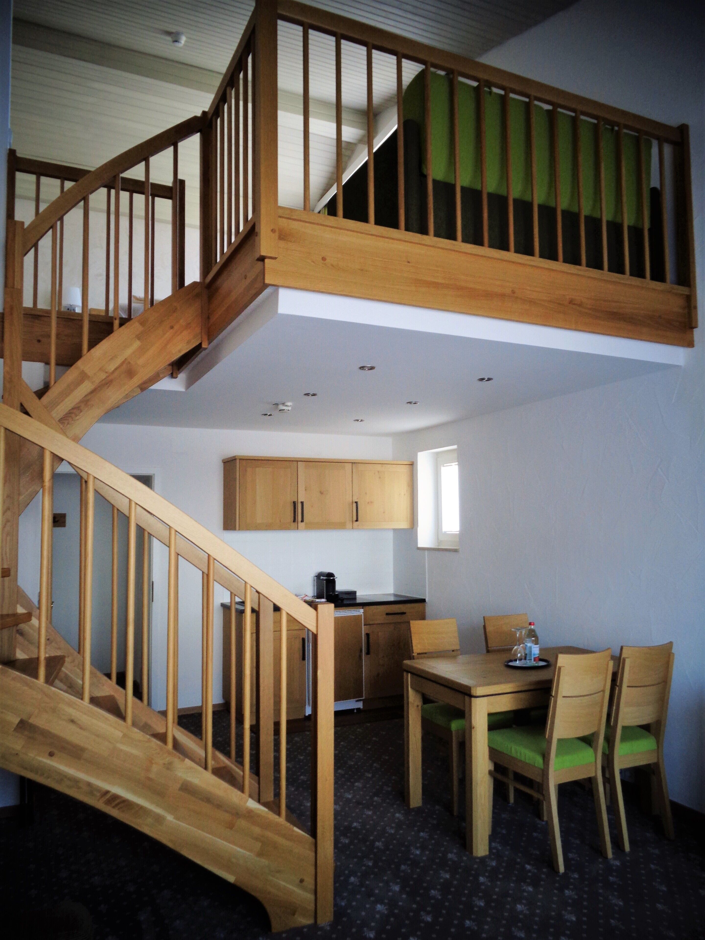 A multi-level wooden staircase connecting two floors, a dining area with a table and chairs, and a kitchen area with cabinets and a coffee maker.
