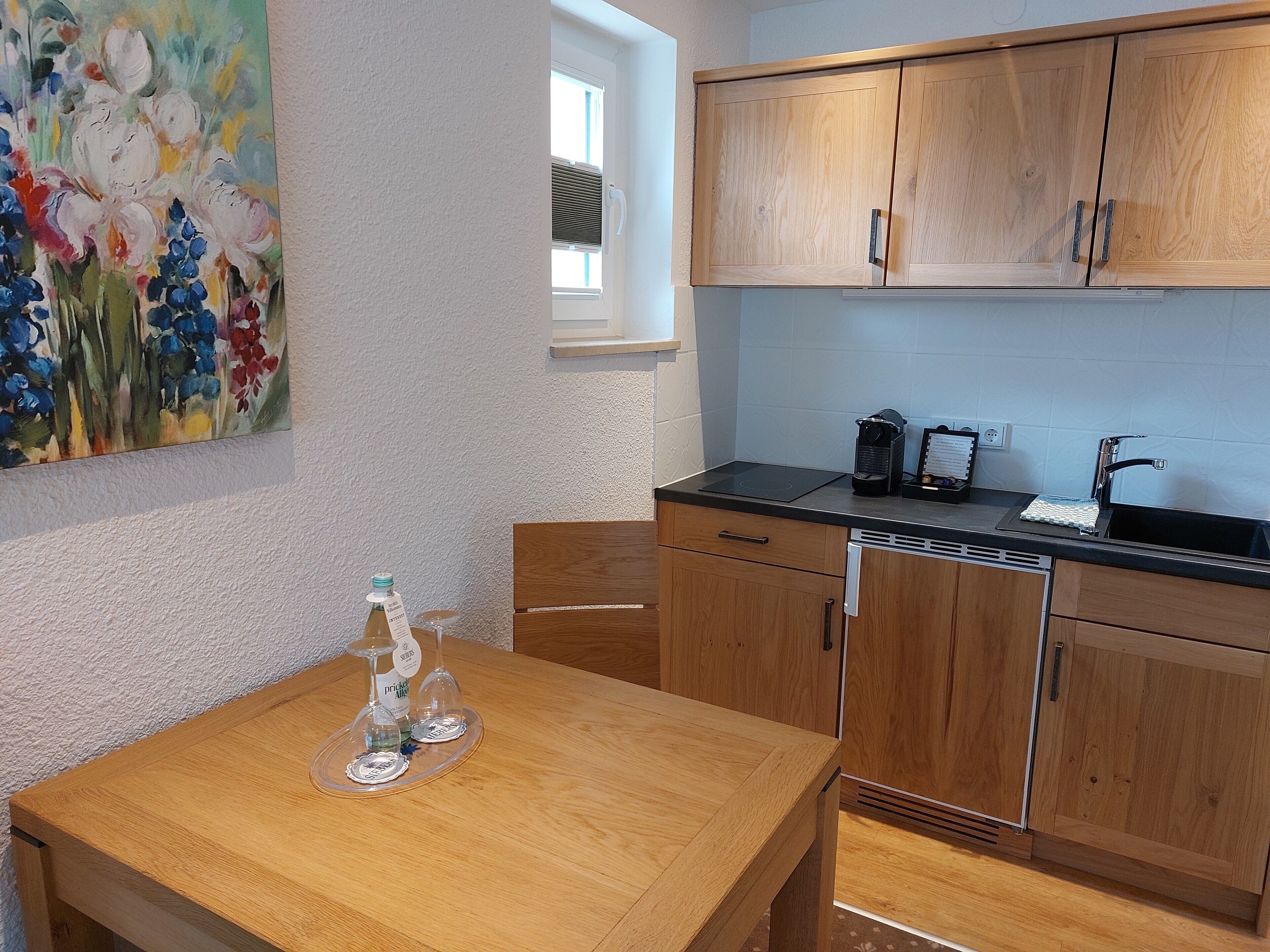 A small kitchen with wooden cabinets, a black countertop, and a window. There is a painting on the wall and a bottle of wine on the table.