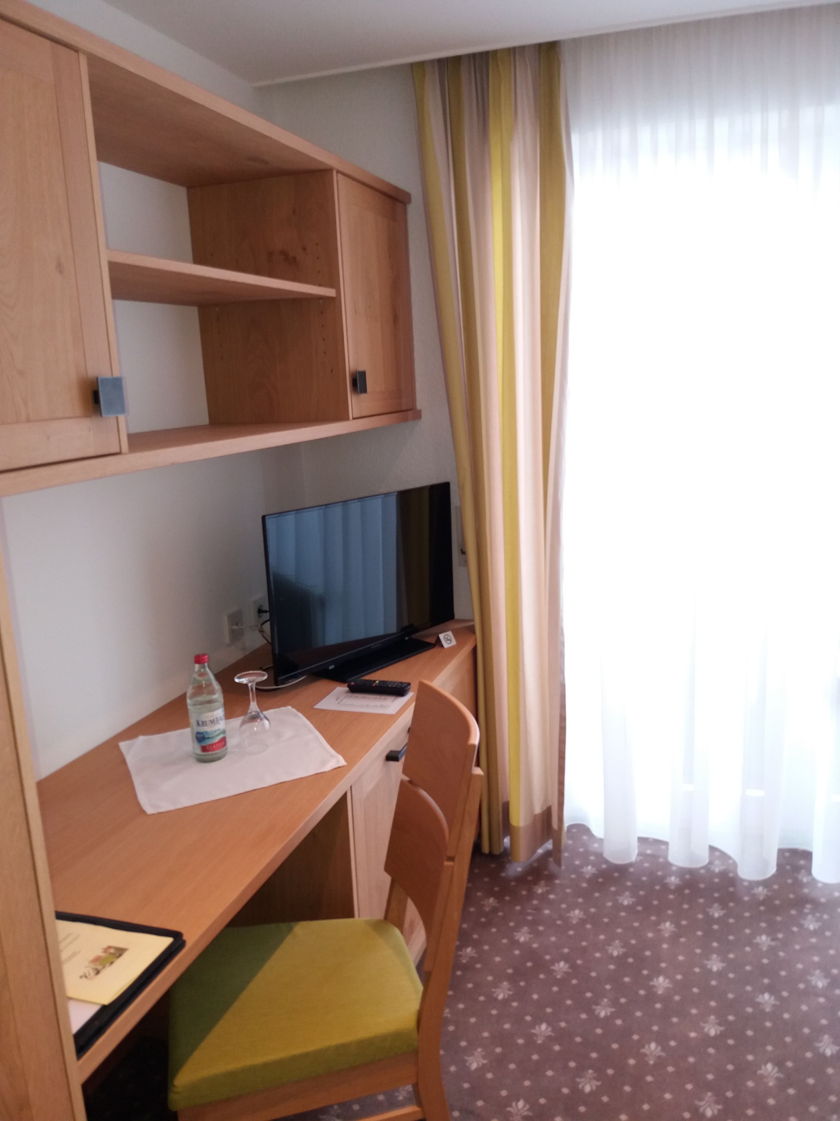 Habitación de hotel con un escritorio de madera, un televisor, una botella de agua, un vaso y una silla con cojín verde.
