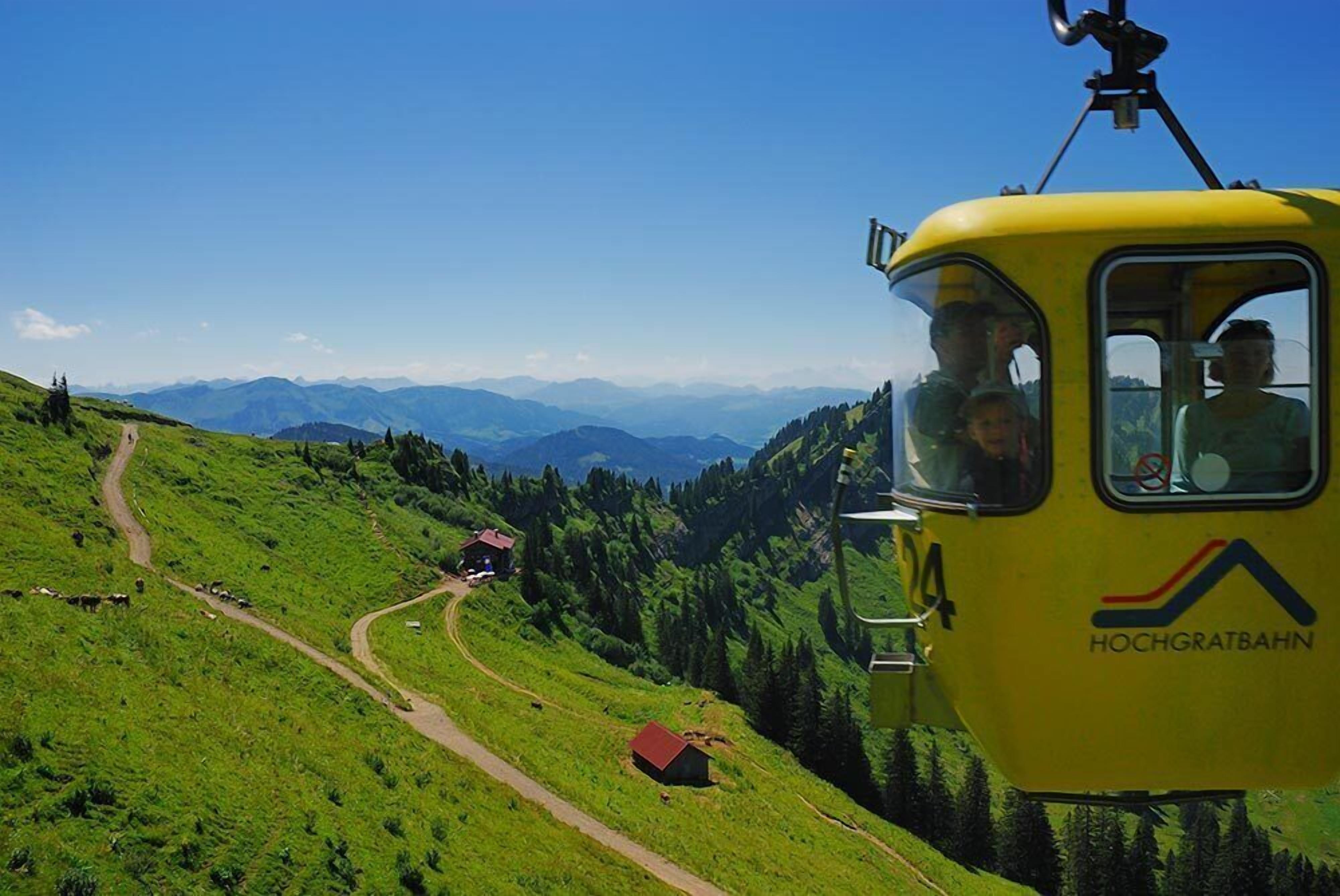 En gul linbana med nummer 24 och texten ”HOCHGRATBAHN” på sidan, som färdas längs en vajer uppför en grönskande sluttning under en klar himmel med avlägsna berg i bakgrunden.