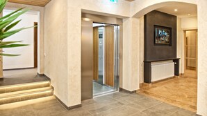 A modern lobby with a glass elevator, a framed artwork on the wall, and a staircase with illuminated steps. -  Interest Vitalhotel (Oberstaufen)
