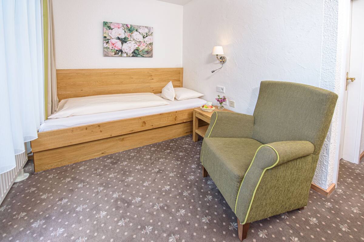 A single bed with a wooden headboard, a green armchair, a small table with a vase of flowers, and a wall-mounted lamp.
