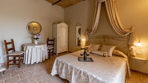A bedroom with a large bed, a nightstand with a lamp, a wardrobe, a round mirror, and a small table with a vase of flowers. - Wine Resort Colsereno (Montalcino)