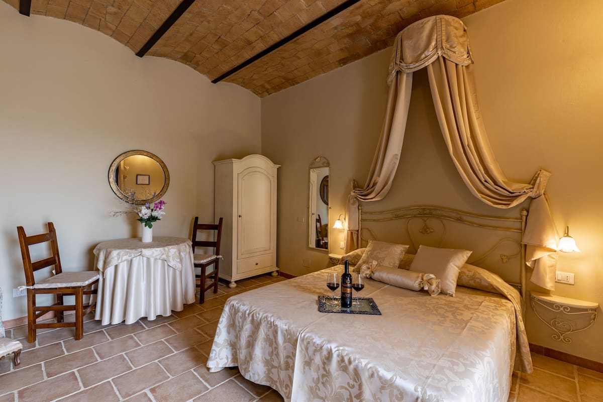 Une chambre avec un grand lit, une table de chevet avec une lampe, une armoire, un miroir rond et une petite table sur laquelle est posé un vase de fleurs.