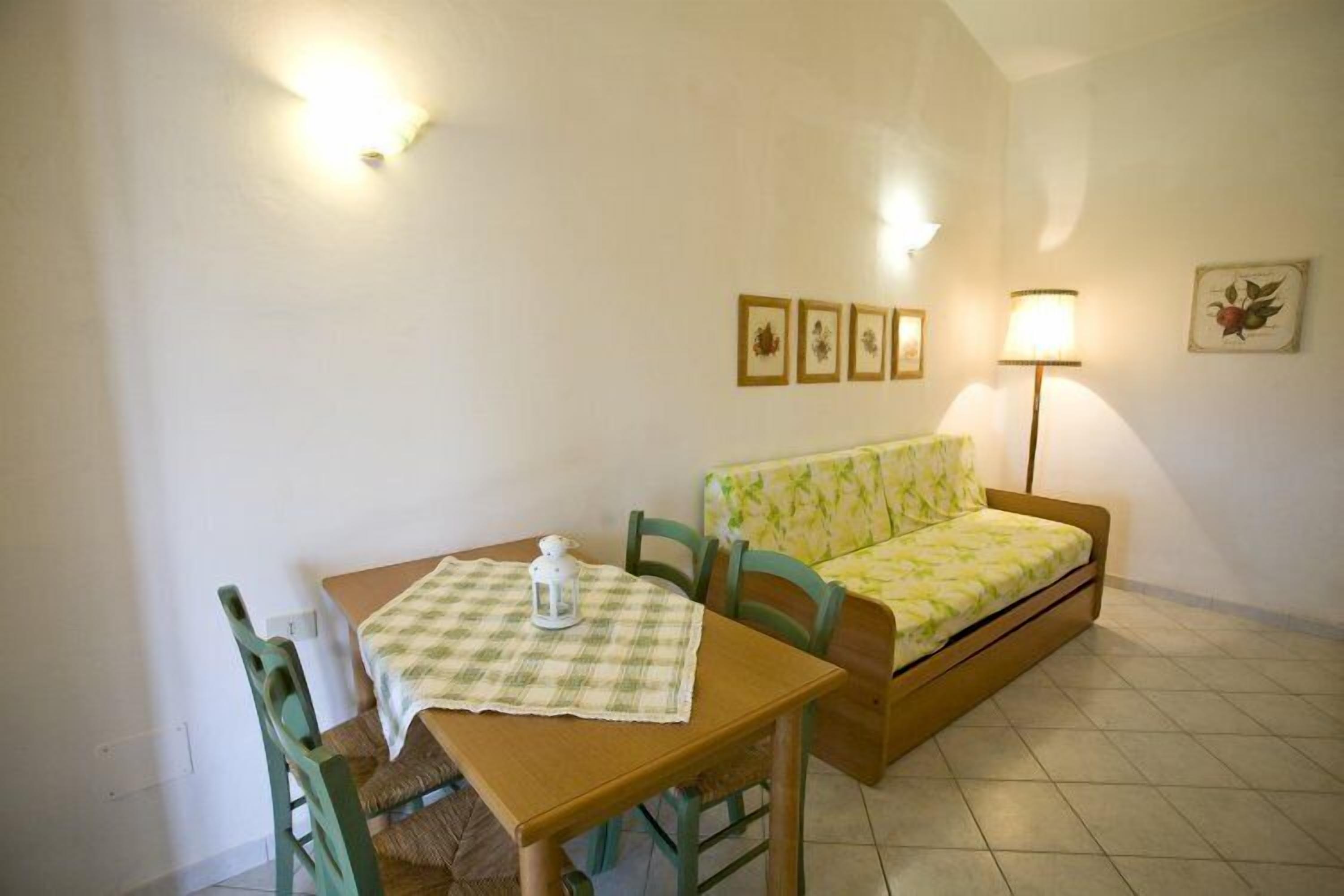 Une petite pièce chaleureuse et accueillante, avec une table en bois, une chaise verte, un canapé et une lampe. Trois tableaux encadrés ornent le mur.