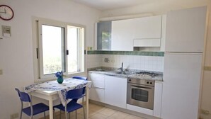 A compact kitchen with white cabinets, a gas stove, and a sink. There's a dining table with blue chairs and a vase of flowers. - Appartamenti Casa Serena (Capoliveri)