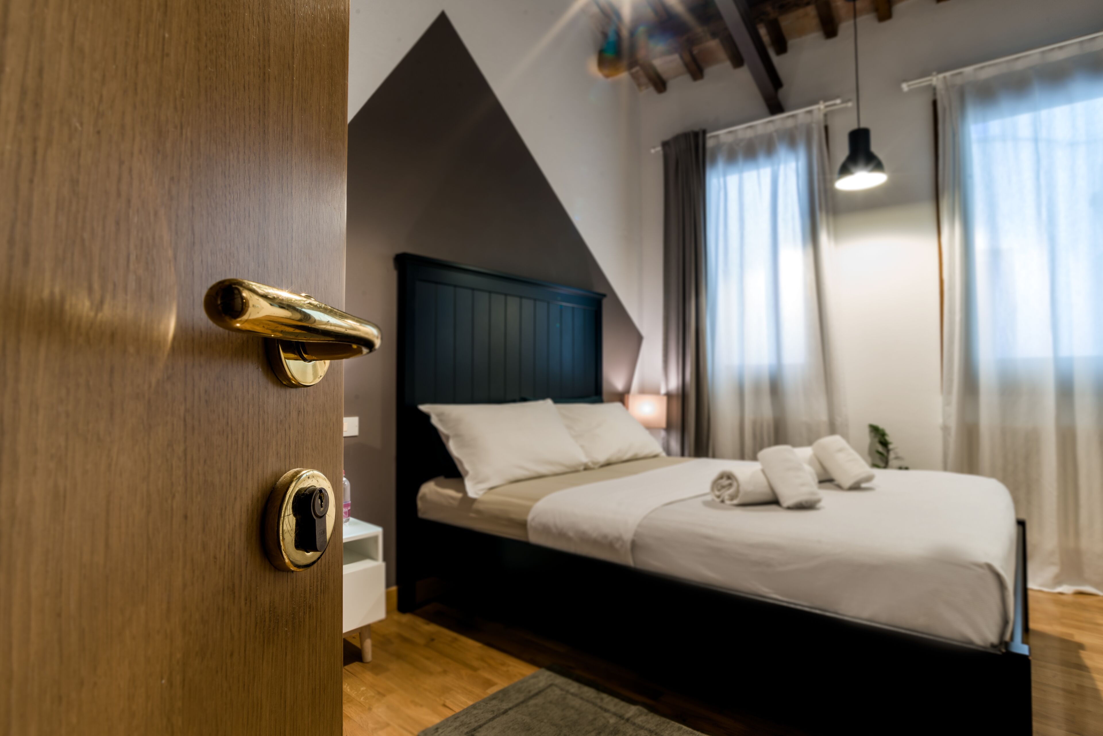 Une chambre avec une porte en bois, un lit avec une literie blanche, une tête de lit sombre et un luminaire suspendu.