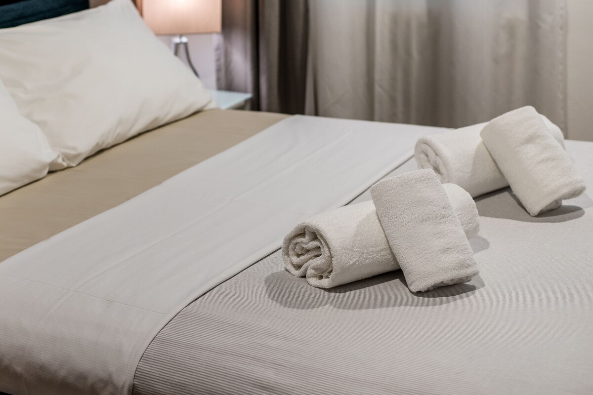 A neatly made bed with white linens, a beige blanket, and two rolled towels.