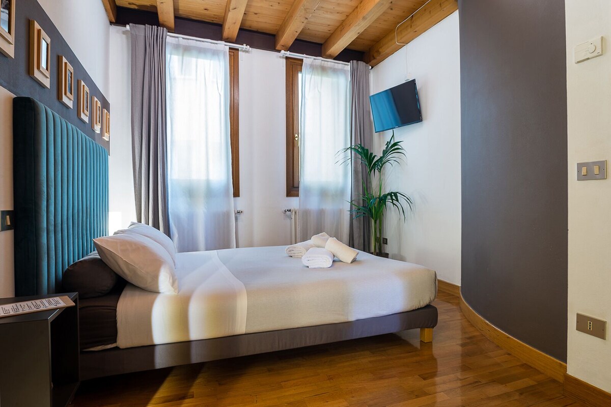 A modern hotel room with a large bed, wooden flooring, a flat-screen TV, and a potted plant.