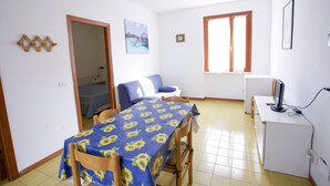 A small, bright room with a dining table covered in a sunflower tablecloth, a sofa, a desk with a computer, and a window with wooden blinds.