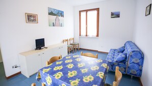 A room with a dining table covered with a sunflower tablecloth, a sofa, a television on a cabinet, and a window with curtains.