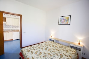 A bedroom with a bed, two bedside tables with lamps, a wall-mounted picture, and a kitchen area with cabinets and a sink. - Appartamenti La Baia Bagnaia (Portoferraio)