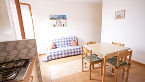 A compact kitchen with a gas stove, a dining table with four chairs, and a blue and white checkered sofa with a cushion.