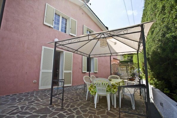 A patio with a table and chairs, a pergola, and a pink building with shutters. - Appartamenti Loredana (Capoliveri)