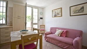 A room with a wooden dining table, a pink sofa, and a window with a view of greenery. - Appartamenti Loredana (Capoliveri)