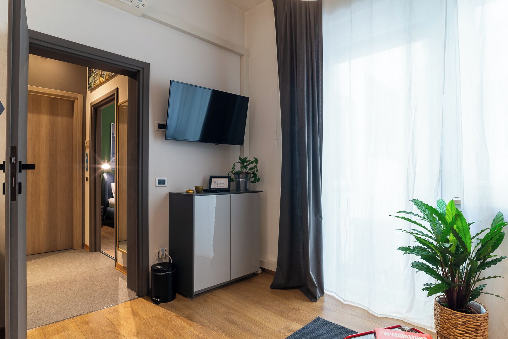 Une chambre d’hôtel moderne équipée d’un téléviseur à écran plat fixé au mur, d’une armoire en bois, d’une plante en pot et d’une grande fenêtre avec des rideaux.