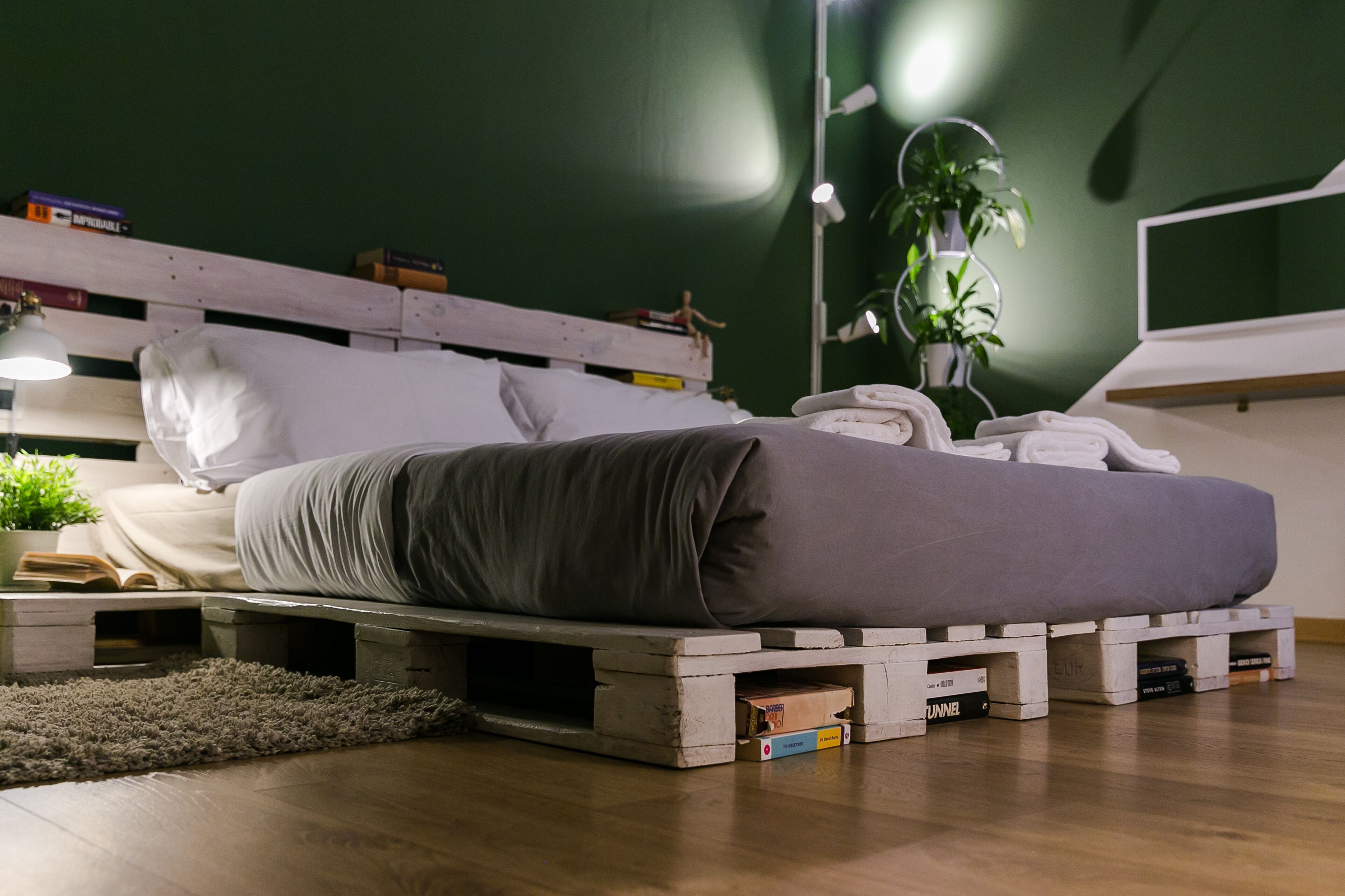 Un lit avec un matelas gris posé sur une structure en bois, une lampe de chevet, une plante en pot et une table de chevet avec des livres.