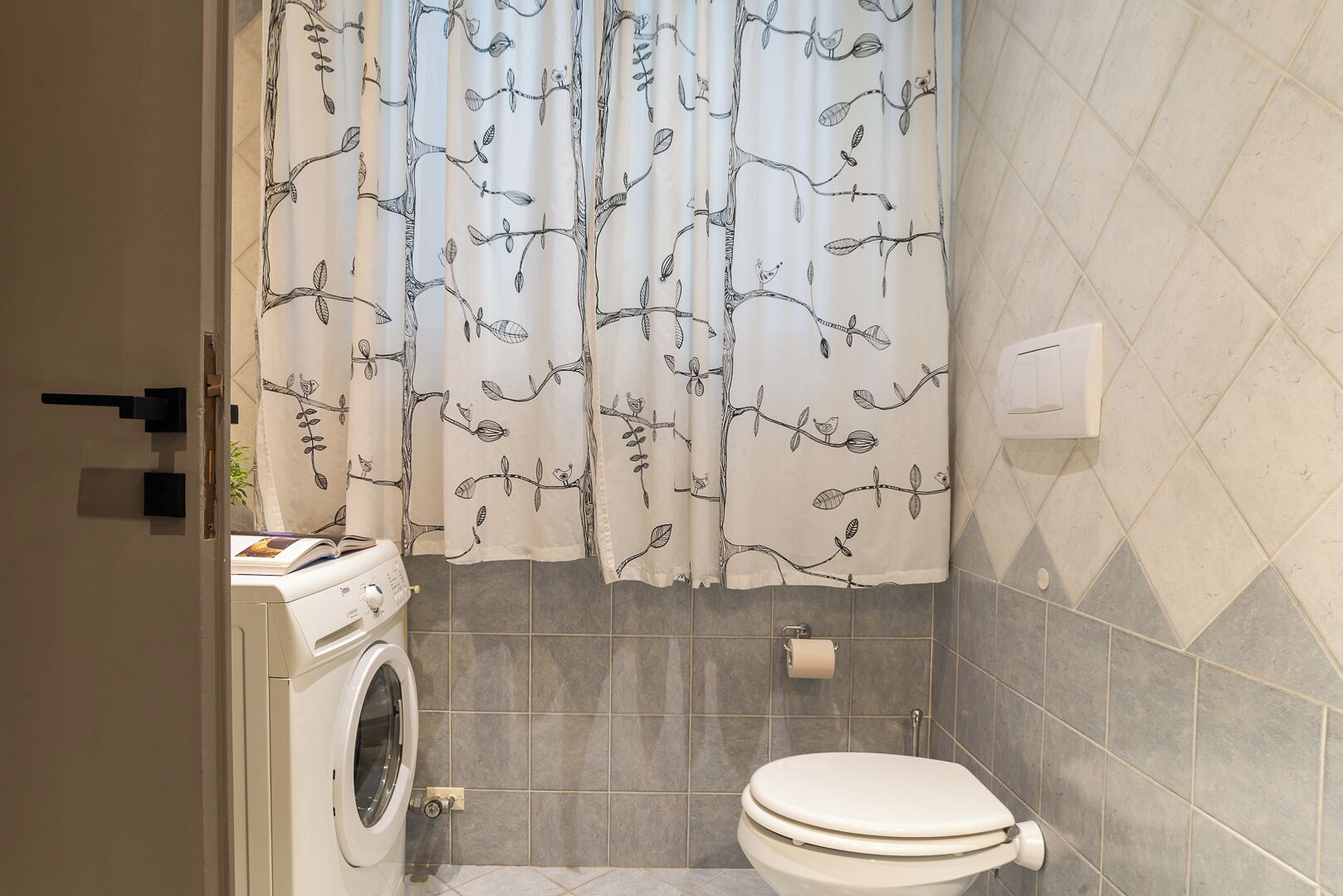 Une salle de bain équipée d’un lave-linge, de toilettes et d’un rideau de douche orné d’un motif représentant une branche d’arbre.