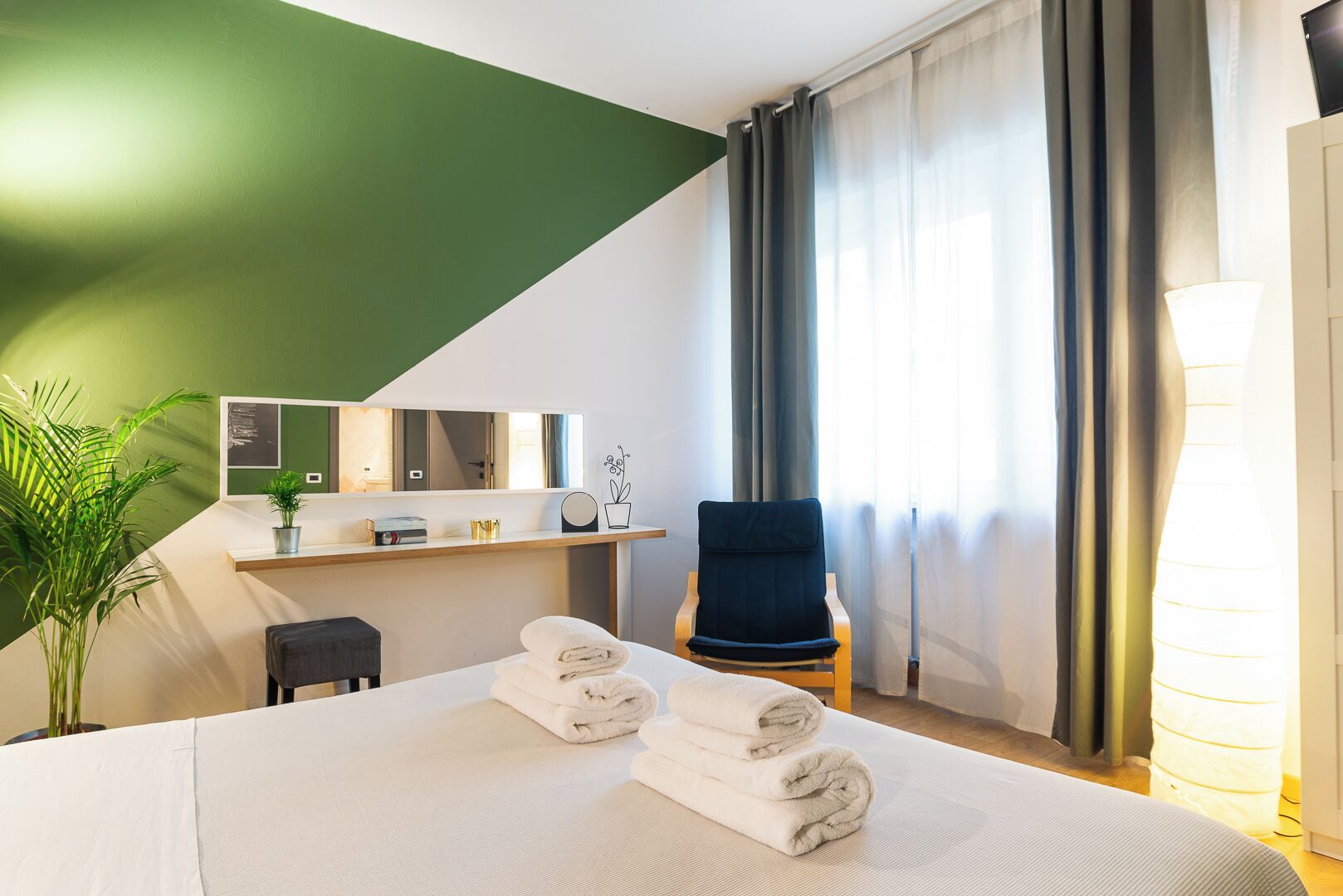 Una camera d'albergo con un letto, una sedia, una scrivania con uno specchio e una pianta in vaso.