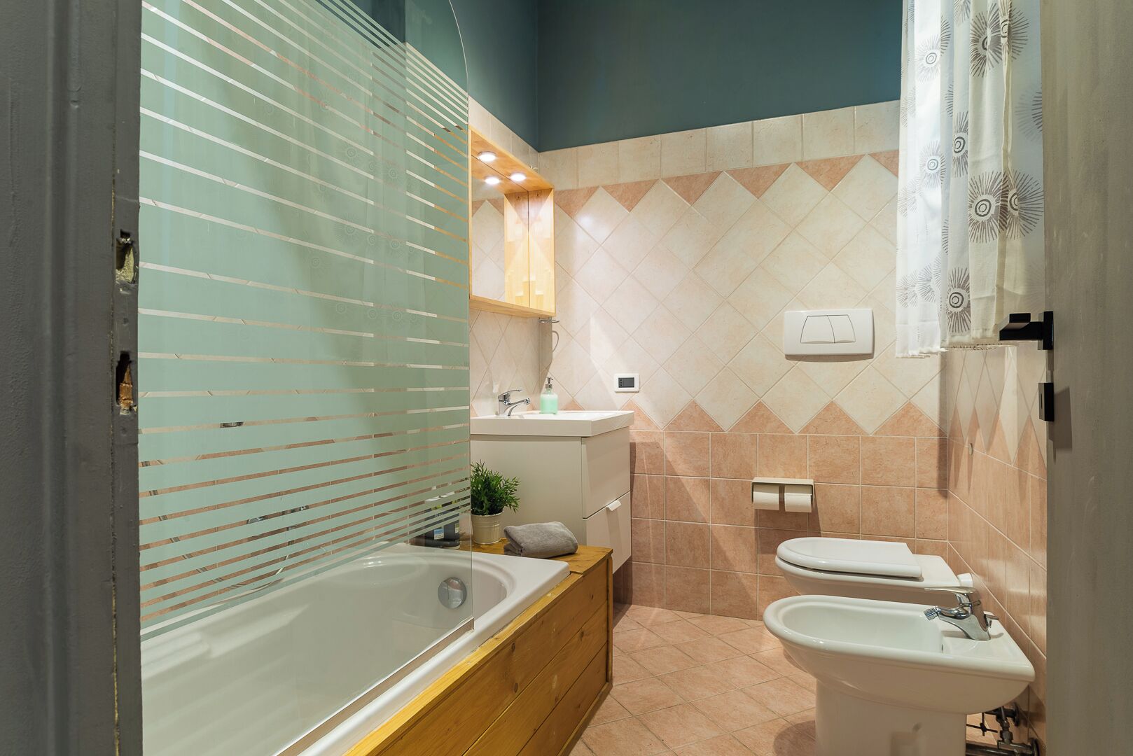 Un baño moderno con una bañera, un lavabo y un inodoro. Hay un mueble de tocador de madera, una planta en maceta y un perchero para toallas.