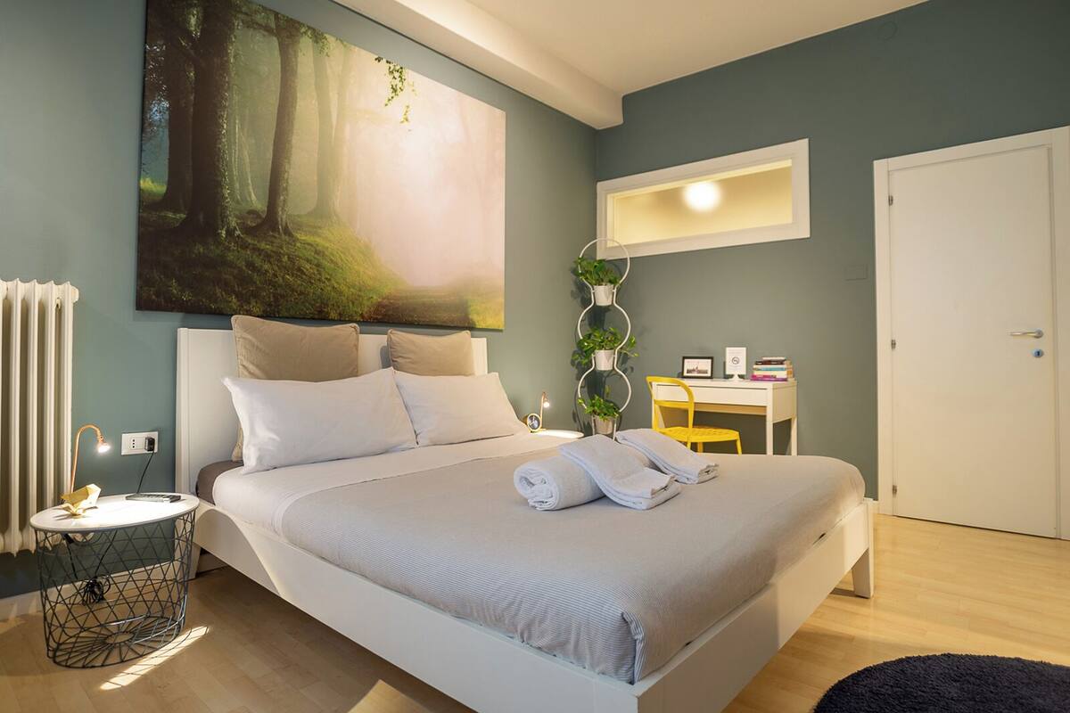 Une chambre bien rangée, avec un grand lit, un bureau, une chaise et un grand tableau accroché au mur.