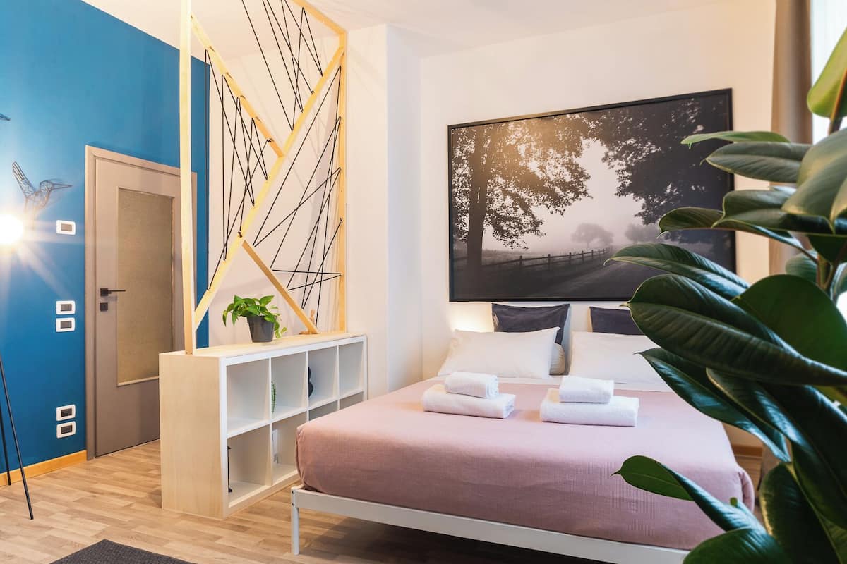 Une chambre moderne avec un lit, une grande fenêtre, une œuvre d’art accrochée au mur et une plante.