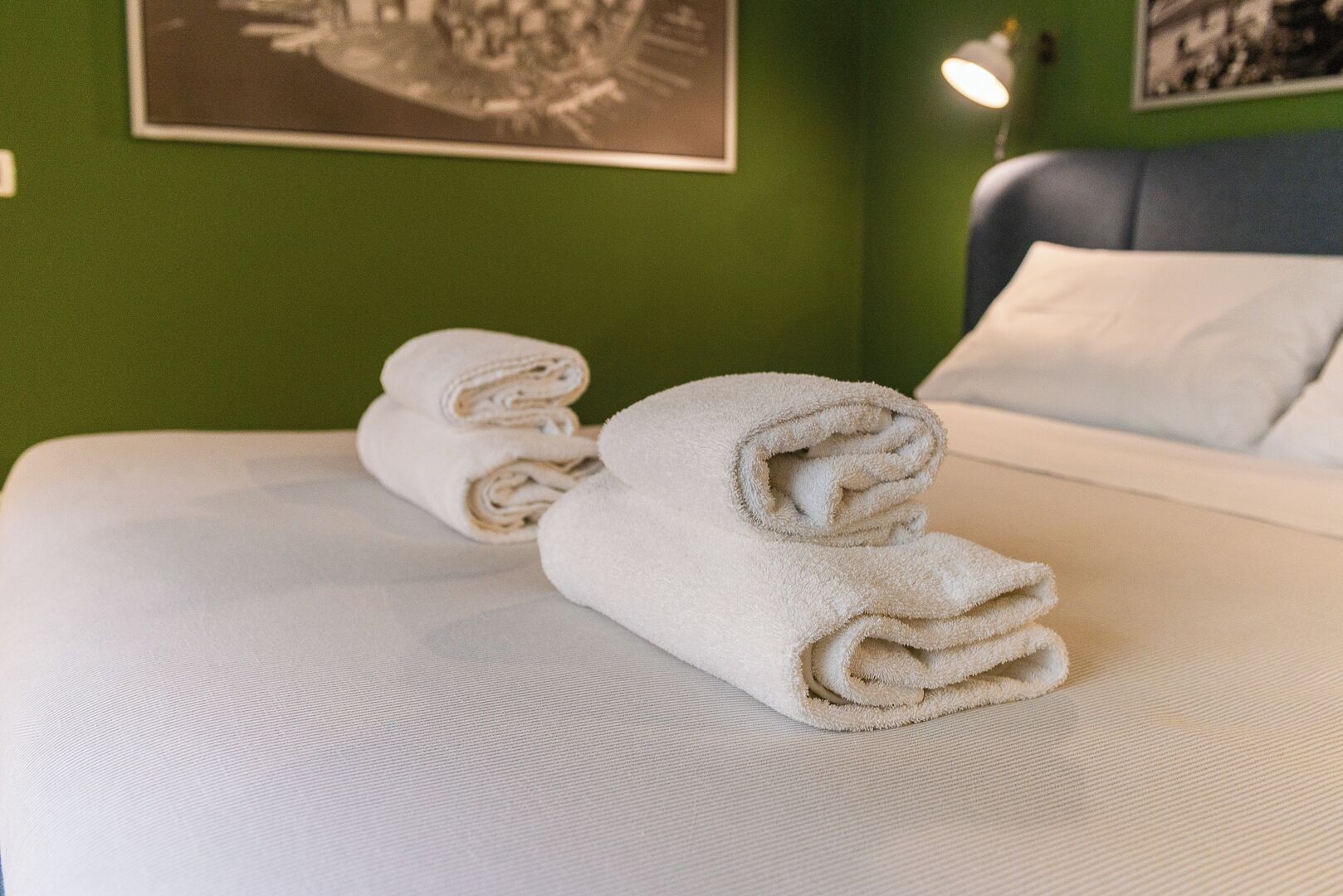 Cama bien hecha con toallas blancas enrolladas y una pared verde con una fotografía blanco y negro.