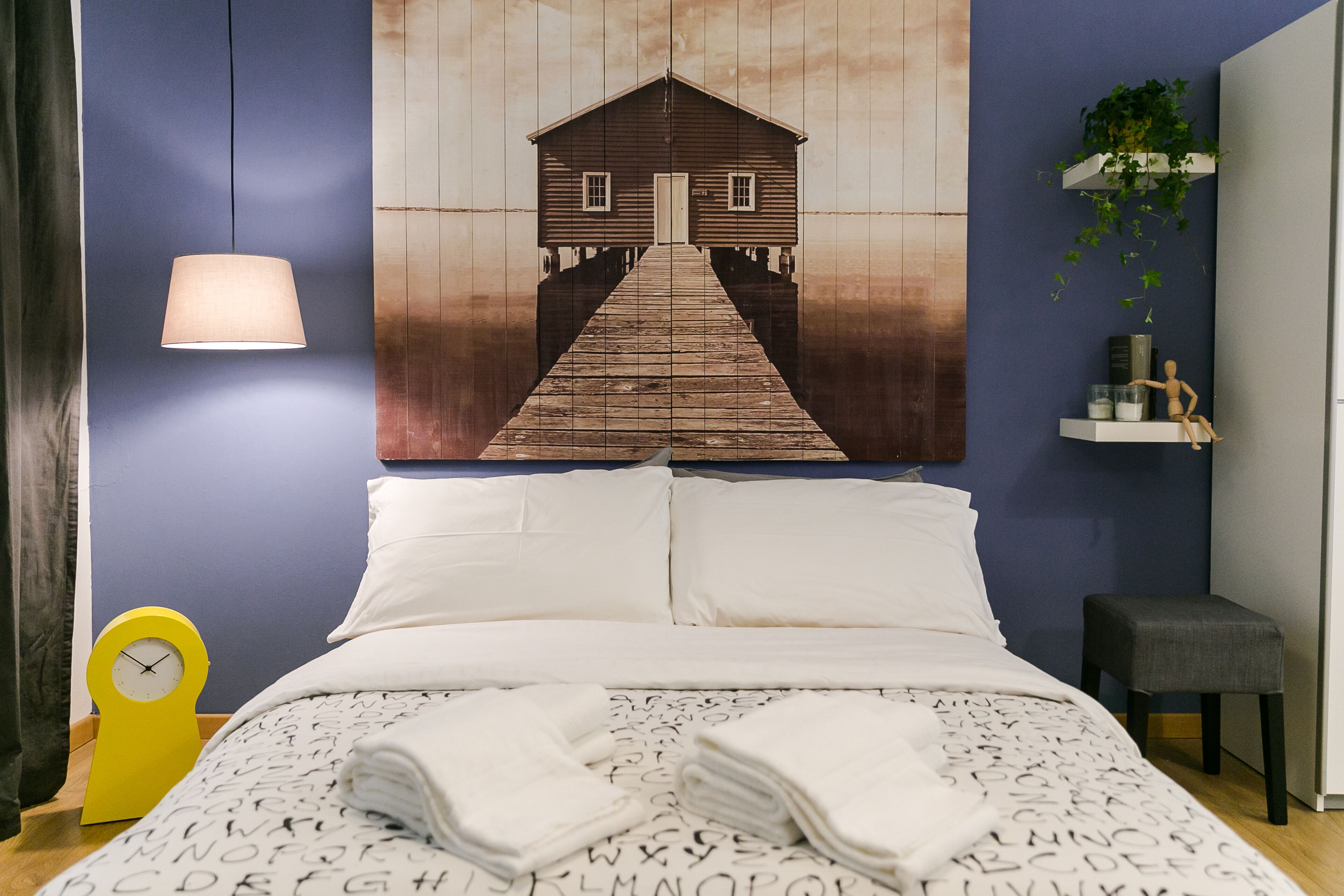 A bedroom with a bed, pillows, a bedside table with a lamp, a clock, and a wall-mounted shelf with a plant.