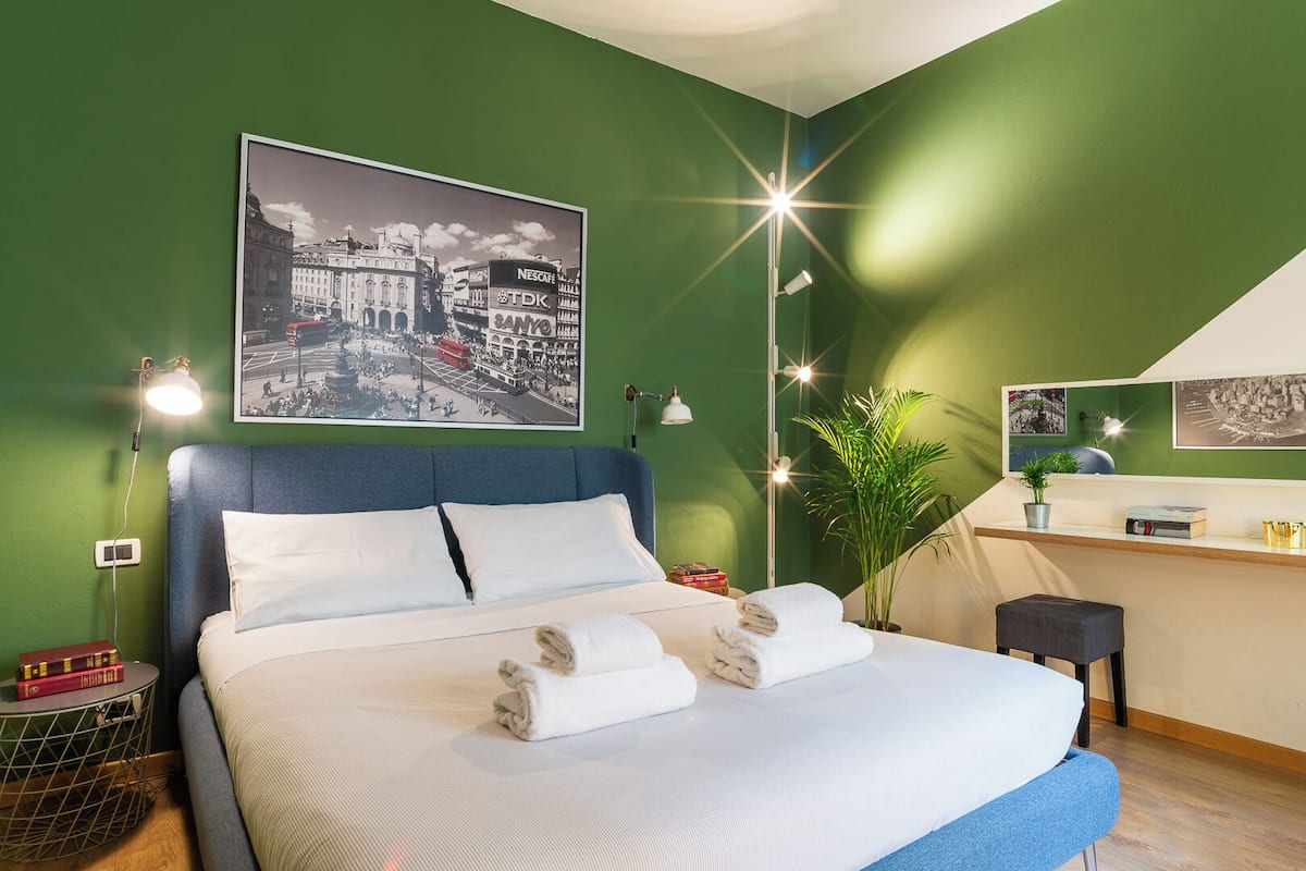 Une chambre d’hôtel avec un grand lit, des murs verts, une affiche représentant un paysage urbain, un petit bureau et une plante en pot.