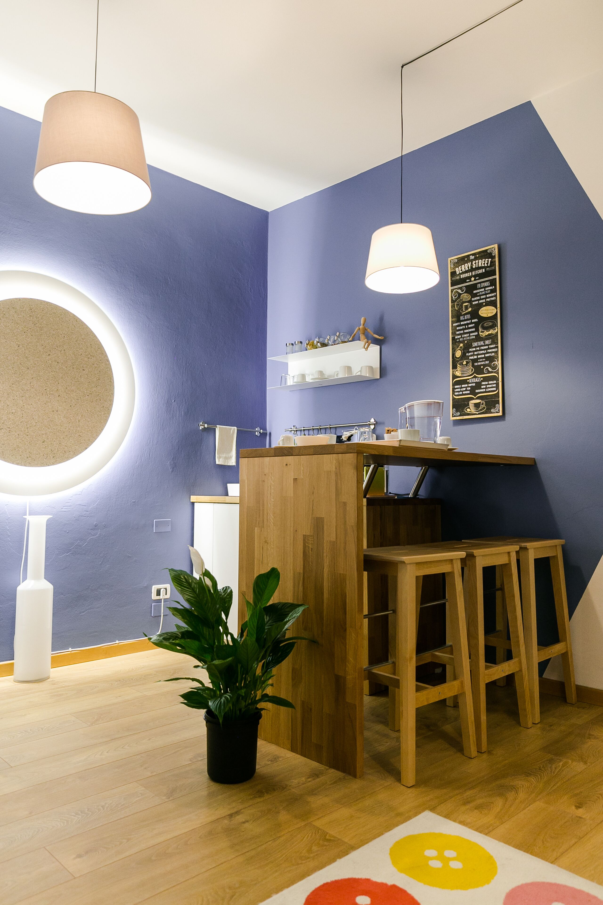A modern kitchen with a wooden counter, bar stools, a hanging light fixture, and a potted plant.