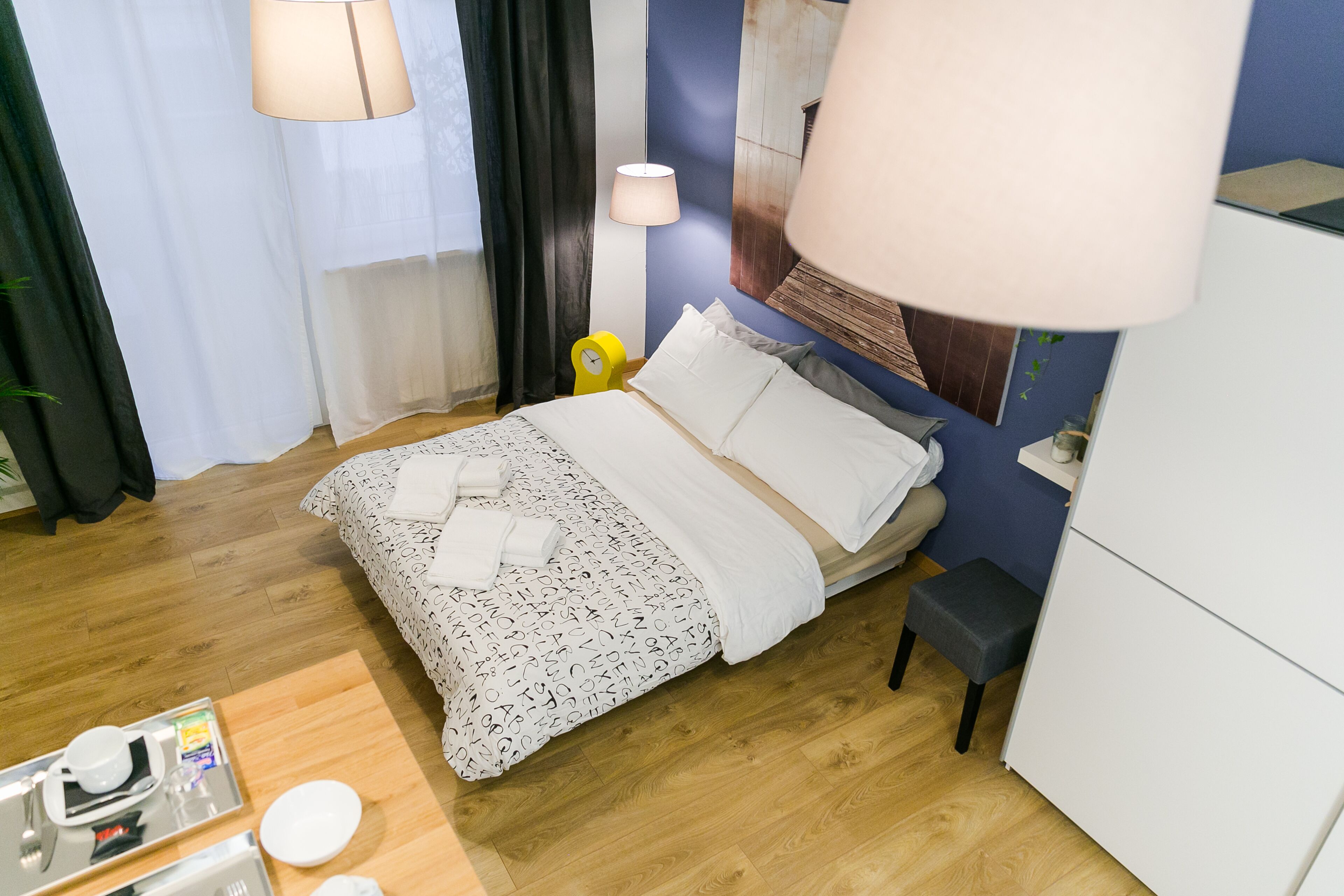 Une chambre d’hôtel bien rangée, avec un lit, une table de chevet, une lampe et une petite table sur laquelle se trouve un plateau avec des articles pour le petit-déjeuner.