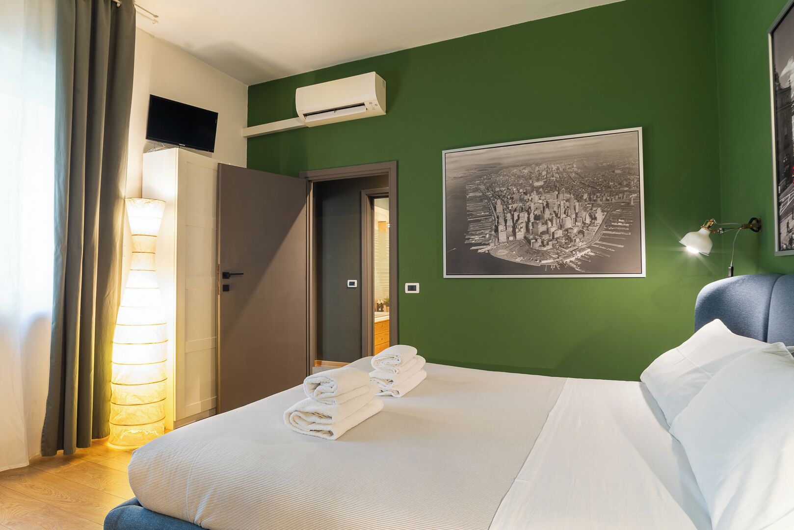 Habitación de hotel con cama, televisión de pantalla plana, aire acondicionado, cuadro enmarco con vista urbana y una lámpara.