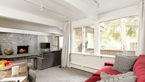Ein modernes Wohnzimmer mit Kamin aus Stein, einem roten Sofa und einem großen Fenster mit Blick auf Bäume.