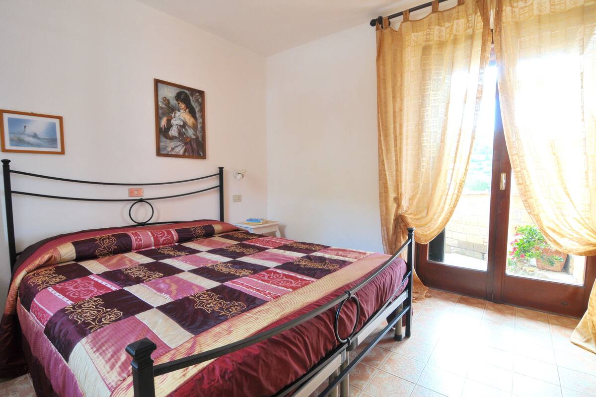 A bedroom with a bed, a nightstand, a window with curtains, and a wall with two framed pictures.