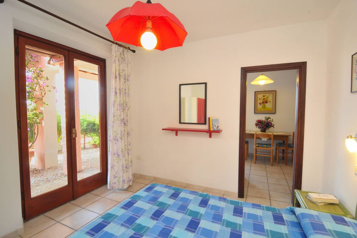 A bedroom with a checkered bedspread, a red umbrella light fixture, a mirror, and a small table with a book.