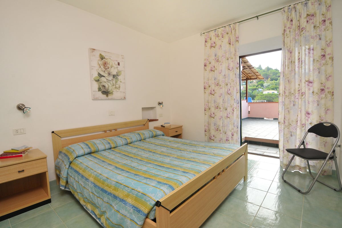 A bedroom with a wooden bed, a nightstand, a chair, and a window with curtains.