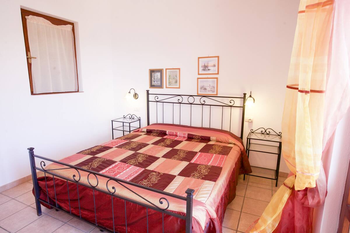 A neatly made bed with a patterned bedspread, a bedside table with a lamp, and a wall-mounted picture frame.