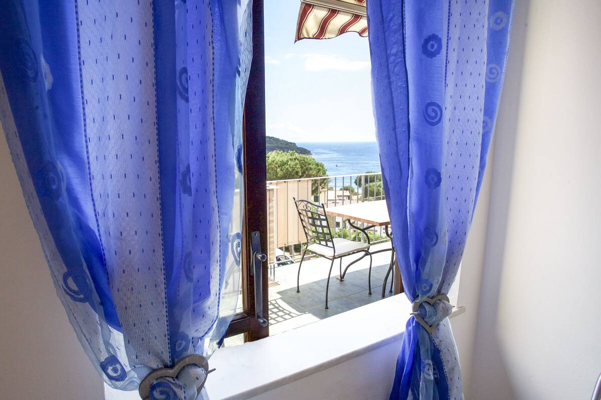 A balcony with a table and chairs, overlooking a scenic view of the sea and hills.