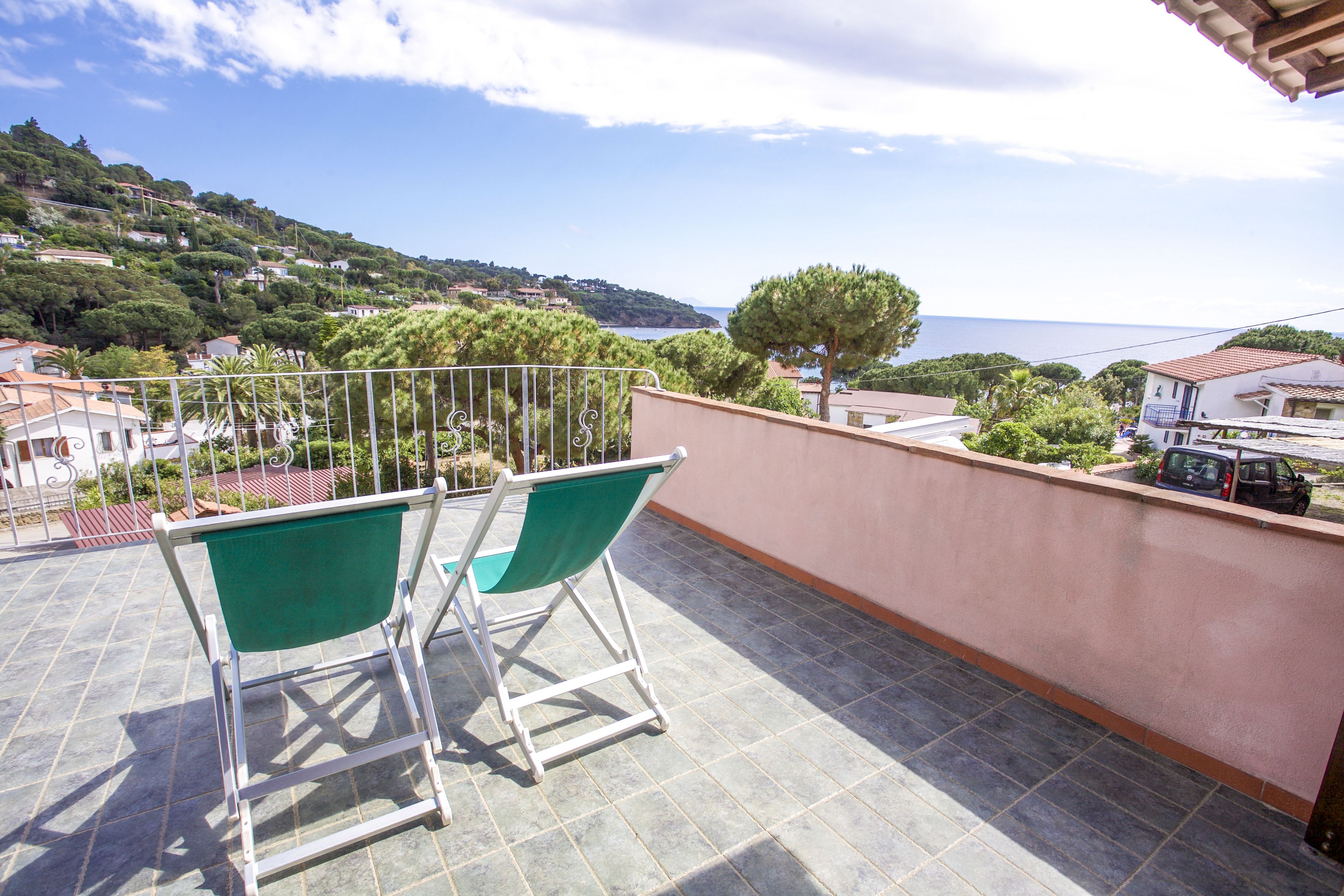 Un balcone con due sedie verdi, vista su una cittadina costiera e cielo sereno.
