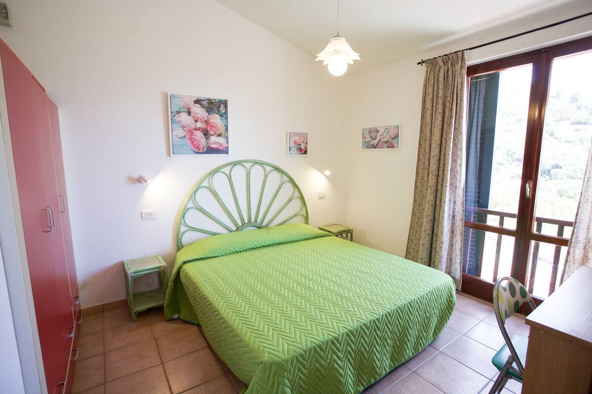 A bedroom with a green bed, a small bedside table, a chair, and a window with curtains.