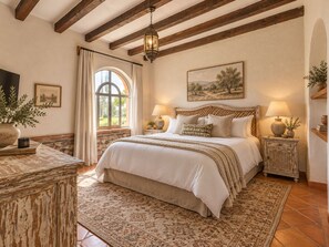 Un dormitorio con una cama grande, vigas de madera, pared de piedra, una ventana con cortinas y un cuadro en la pared.