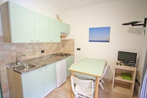 A compact kitchen with a sink, stove, and white cabinets. There is a small dining table with chairs and a television on a wooden stand.